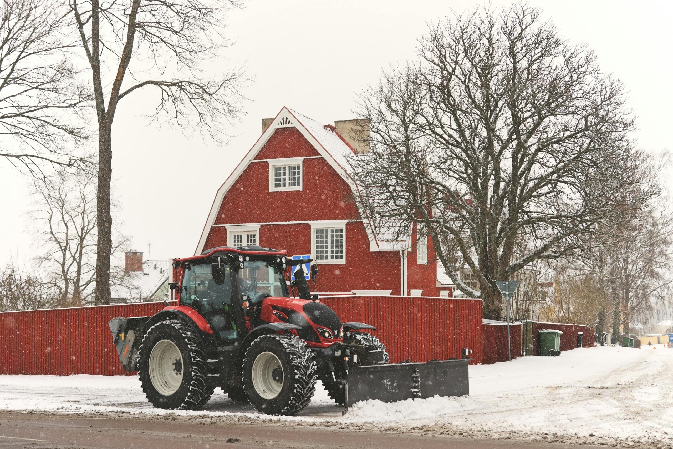 A red tractor plowing snow near a traditional red house in Jönköping, Sweden.